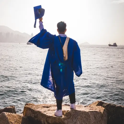 a person in graduation apparel, facing a lake and holding their graduation cap in the air