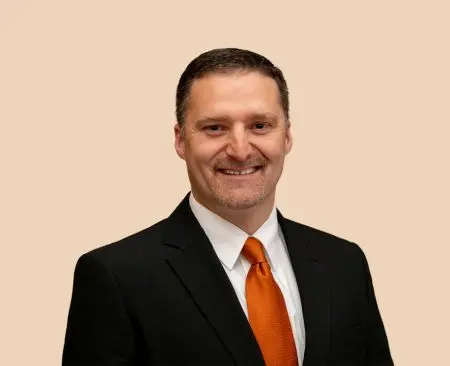 man smiling wearing black suit jacket over a white button-up shirt with orange tie